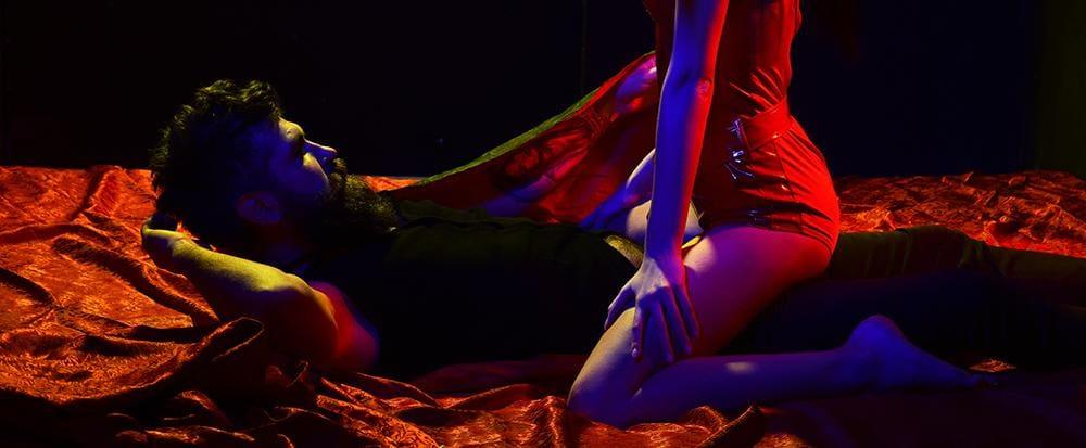 Man lying on red satin sheets with a woman in a red outfit kneeling over him in low blue and red lighting