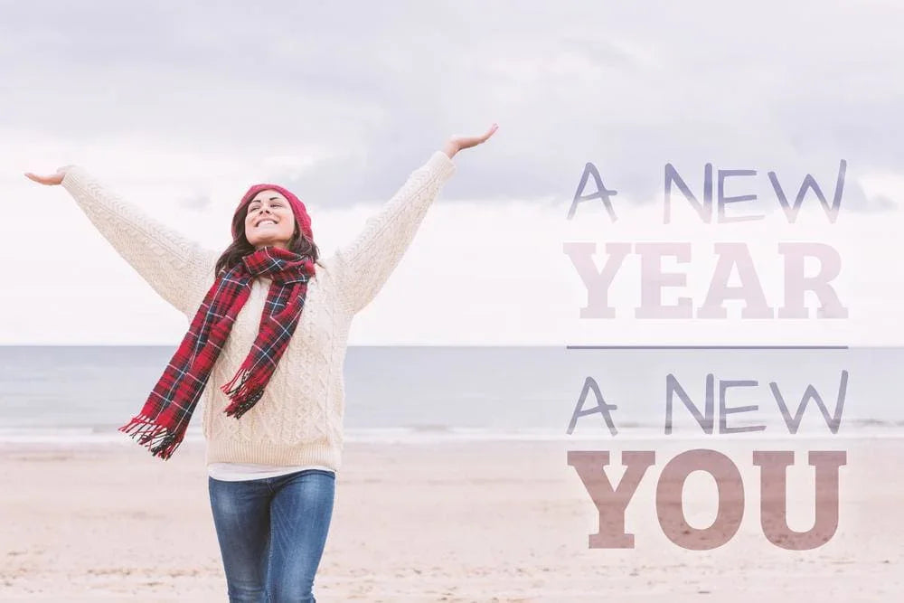 Happy woman wearing cozy sweater, red scarf, and beanie on beach with inspirational New Year message