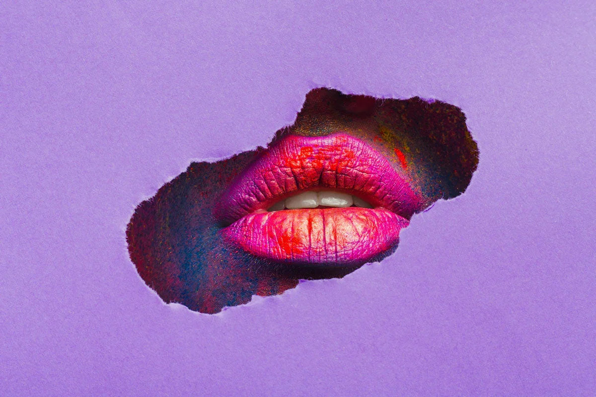 Close-up of vibrant pink lips with red and orange paint, seen through torn purple paper